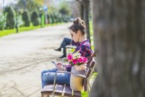 Vista lateral de una joven mujer hipster sentada en un banco del parque relajándose en un día soleado mientras usa un teléfono móvil - foto de stock
