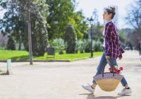 Vista lateral de uma jovem hipster segurando uma cesta de vime enquanto caminhava em um parque em dia ensolarado — Fotografia de Stock