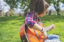 Vista frontal de uma jovem hipster usando óculos de sol, sentada na grama em um parque enquanto aprecia tocar guitarra — Fotografia de Stock