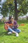 Vista frontale di una giovane hipster che indossa occhiali da sole, seduta sull'erba in un parco mentre si diverte a suonare la chitarra — Foto stock