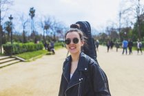 Vorderansicht einer jungen lächelnden Hipsterfrau, die an sonnigen Tagen in einem Park spaziert, während sie eine Gitarre auf dem Rücken trägt — Stockfoto