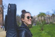 Vista lateral de uma jovem mulher hipster sorridente andando em um parque em dia ensolarado enquanto carrega uma guitarra nas costas — Fotografia de Stock