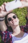 Vista acima de uma jovem mulher hipster sorridente deitada na grama em um dia ensolarado em um parque enquanto toma uma selfie com um telefone celular — Fotografia de Stock