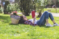 Vue latérale d'une femme heureuse hipster allongée sur l'herbe dans une journée ensoleillée au parc tout en lisant un livre rouge — Photo de stock