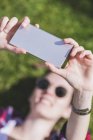 Above view of a young smiling hipster woman lying on grass in a sunny day at a park while taking a selfie with a mobile phone — Stock Photo