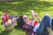 Vista lateral de uma mulher feliz hipster deitado na grama em dia ensolarado no parque, enquanto usando um telefone móvel — Fotografia de Stock