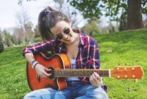 Вид спереду молода жінка-хіппер в сонцезахисних окулярах, сидить на траві в парку, насолоджуючись грою на гітарі — стокове фото