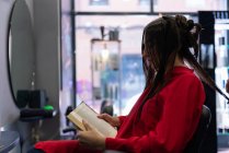 Side view of attractive young lady reading volume and sitting on chair with beautiful hairstyle in hairdressing salon — Stock Photo