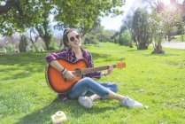Front view of a young hipster woman wearing sunglasses, sitting on grass in a park while enjoying playing guitar — Stock Photo