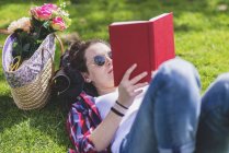 Вид сбоку счастливой женщины-хипстера, лежащей на траве в солнечный день в парке во время чтения красной книги — стоковое фото
