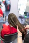 Cortar las manos de estilista peinando el pelo de la mujer morena sentada en sillas en el salón de peluquería - foto de stock