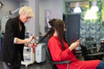 Mujer joven con teléfono móvil y sentado en la silla con hermoso peinado en el salón de peluquería - foto de stock