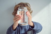 Carino bambino sorridente e in possesso di vaso di vetro con acqua pulita davanti al viso mentre in piedi contro muro bianco — Foto stock