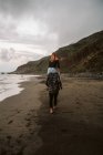 Vista trasera de tipo descalzo dando paseo a cuestas a la señora anónima mientras camina en la orilla del mar arenoso en el día nublado - foto de stock
