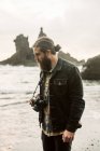 Бородатий чоловік з професійною фотокамерою, дивлячись далеко, стоячи біля моря — стокове фото