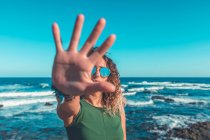 Trendy lady in sunglasses showing stop gesture to camera while standing near beautiful waving sea against cloudless blue sky on sunny day — Stock Photo