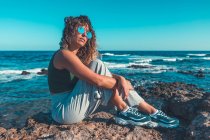 Вид сбоку привлекательной мечтательной женщины в трикотажном наряде и солнцезащитных очках, опускающей колени и отводящей взгляд, сидя на берегу у моря — стоковое фото
