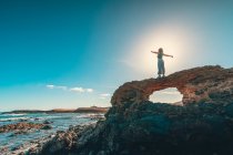 Неузнаваемая женщина с вытянутыми руками, стоящая на грубой скале возле величественного моря против ярко-синего неба в солнечный день — стоковое фото