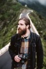 Бородатий стильний хлопець з професійною фотокамерою, дивлячись далеко, стоячи на асфальтній дорозі на гірському хребті — стокове фото