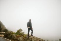 Vista trasera del hombre en traje casual de pie en el acantilado rocoso cerca de la carretera rural en el día de niebla en España - foto de stock