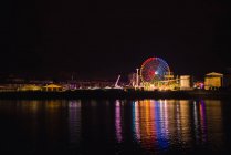 Parque de atracciones brillante con gran rueda de observación en luces de colores que reflejan en el agua del canal de la ciudad en la noche - foto de stock