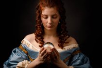 Pretty woman in vintage Victorian dress looking down while holding magical glass ball. — Stock Photo