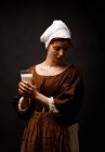 Pretty woman in simple medieval dress holding glass of fresh milk on black background. — Stock Photo