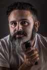 Красивий дорослий чоловік використовує електричну бритву, щоб поголити бороду і дивиться на камеру, стоячи на сірому фоні — стокове фото
