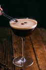 Decorating espresso martini cocktail in glass with coffee beans on wooden table — Stock Photo