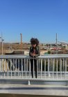 Frau mit Afrohaaren blickt auf ihr Handy, während sie auf einer Brücke in einer Stadt spaziert — Stockfoto
