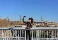 Женщина с афроволосами смотрит в телефон, гуляя по мосту в городе — стоковое фото