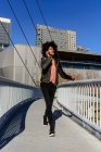 Femme aux cheveux afro regardant son téléphone en marchant sur un pont dans une ville — Photo de stock