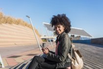 Чорна жінка з афро волоссям сидить на вулиці зі своїм смартфоном у руці, посміхаючись — стокове фото