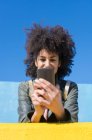 Mujer negra con cabello afro apoyada en paredes de colores brillantes mientras mira su smartphone y toma un café - foto de stock