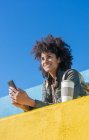 Femme noire aux cheveux afro s'appuyant sur des murs aux couleurs vives tout en regardant son smartphone et en prenant un café — Photo de stock