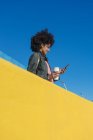 Чорна жінка з афро волоссям, що спирається на яскраво-кольорові стіни, дивлячись на її смартфон і маючи каву — стокове фото