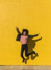 Schwarze Frau mit Afro-Haaren springt vor Freude auf die Straße mit einer gelben Wand im Hintergrund — Stockfoto