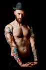 Shirtless bald serious hipster with tattoos on arms and hat looking at camera on black background — Stock Photo