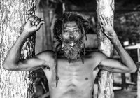 Бородатый афроамериканец с дредами и поднятыми руками курит сигару возле деревьев — стоковое фото