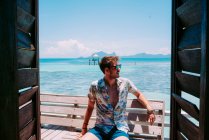 Young guy in sunglasses sitting on seat near blue sea and looking away in Jamaica — Stock Photo