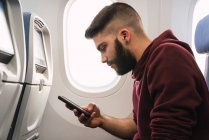Бородатий хлопець використовує смартфон у літаку — стокове фото