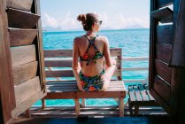 Vista posteriore della signora in costume da bagno e occhiali da sole seduta sul sedile vicino al mare blu in Giamaica — Foto stock