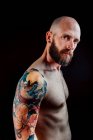 Side view of shirtless bald serious hipster with tattoos on hands looking at camera on black background — Stock Photo