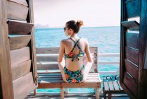 Back view of lady in swimsuit and sunglasses sitting on seat near blue sea in Jamaica — Stock Photo
