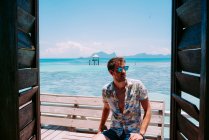 Young guy in sunglasses sitting on seat near blue sea and looking away in Jamaica — Stock Photo