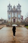 Visão traseira da fêmea com guarda-chuva branco caminhando no pavimento antigo em direção à bela construção da igreja no dia chuvoso no Porto, Portugal — Fotografia de Stock