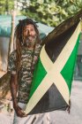 Africano americano barbudo macho com dreadlocks segurando Jamaica bandeira perto da árvore — Fotografia de Stock