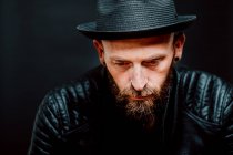 Cool hipster in hat and leather jacket looking down on black background — Stock Photo