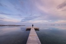 Vista posteriore di una persona irriconoscibile in piedi sul lungo molo di legname vicino all'acqua di mare calma la sera nuvolosa in Spagna — Foto stock