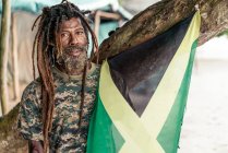 Africano americano barbudo macho com dreadlocks segurando Jamaica bandeira perto da árvore — Fotografia de Stock